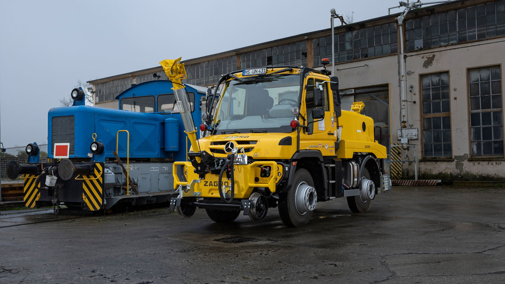 Ein gelber Zweiwege Unimog steht neben einem blauen Lokomotivfahrzeug. Beide Fahrzeuge sind auf einer gepflasterten Fläche platziert, mit einem Gebäude im Hintergrund.