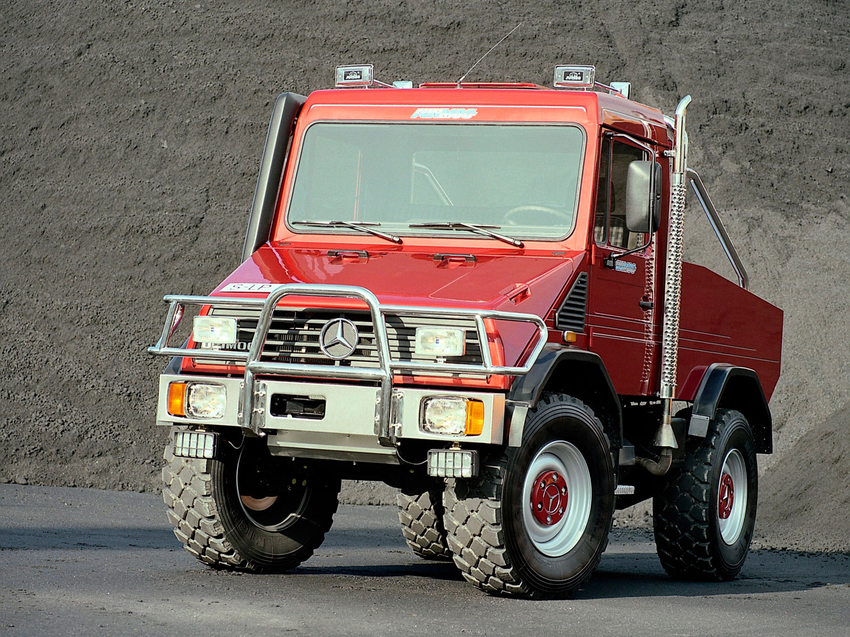 Rode Unimog van Mercedes‑Benz met een extra laadplatform staat op een stoffig oppervlak, ideaal voor offroad- en transporttoepassingen in ruwe omgevingen.