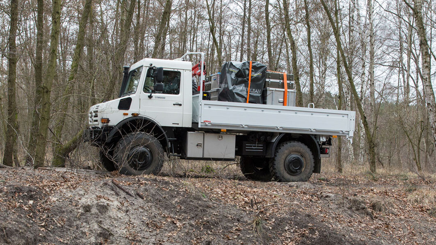 Ein Mercedes-Benz Unimog steht auf einem Erdhang im Wald, mit Ladung auf der Ladefläche, umgeben von Baumstämmen und Laub.