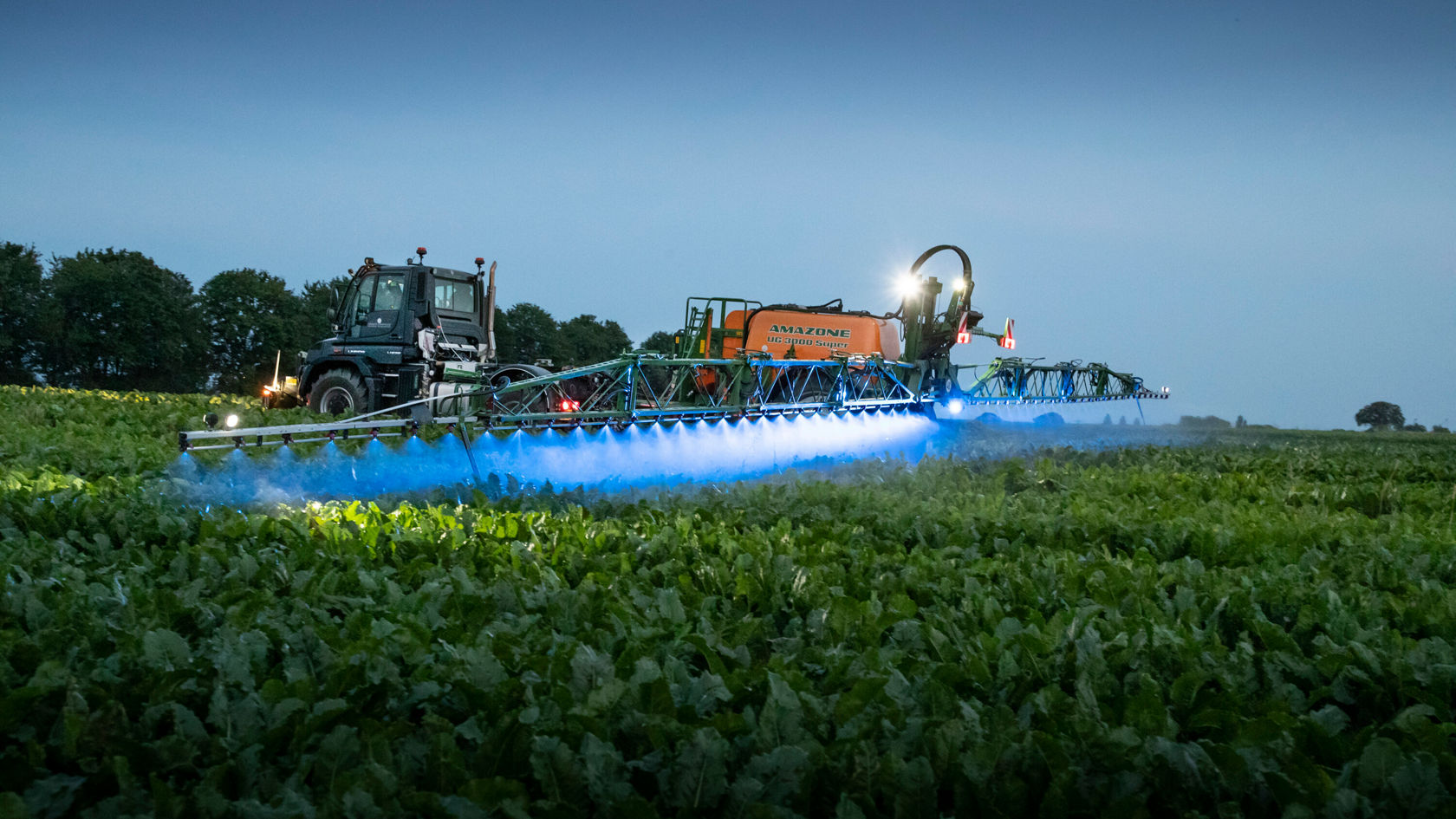 Un tracteur pulvérise des produits phytosanitaires dans un champ au crépuscule. La rampe de pulvérisation émet une lueur bleue et est clairement visible, tandis que les environs sont partiellement à l'ombre.