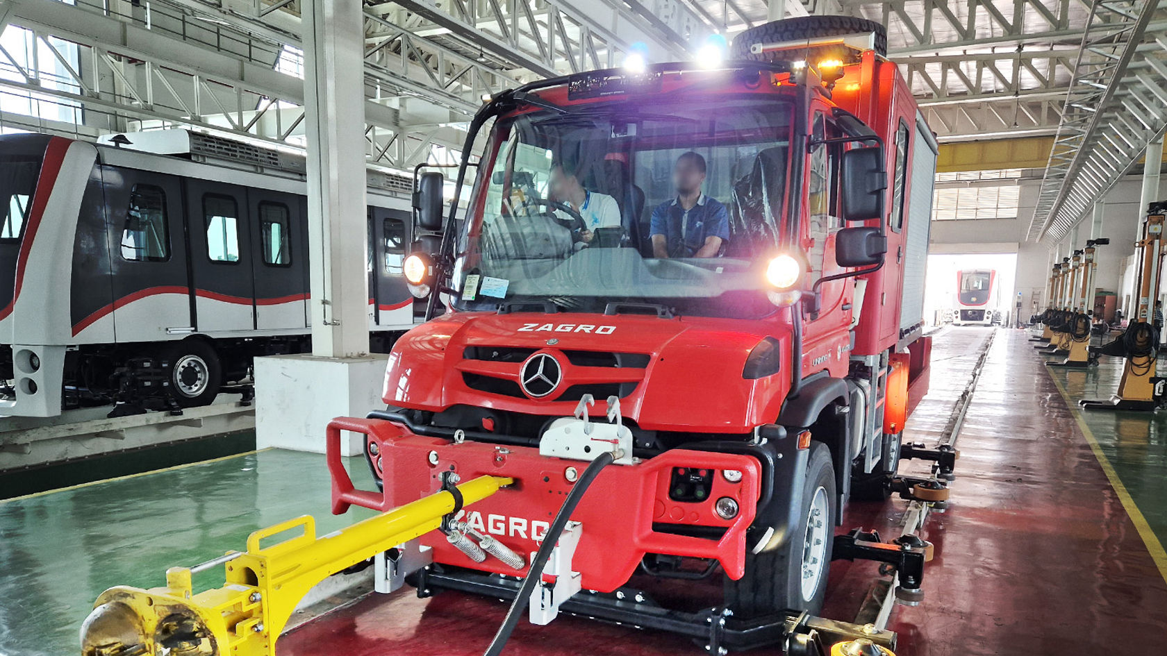 Roter Mercedes-Benz Unimog mit einem gelben Anbaugerät fährt durch eine Halle, während zwei Personen im Führerhaus sitzen.