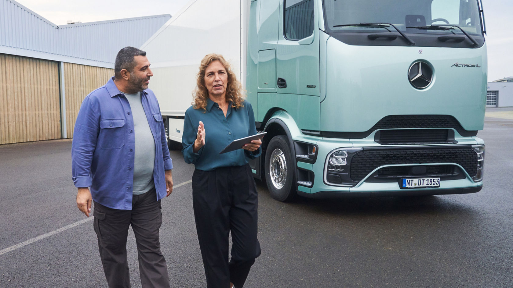 Eine Frau und ein Mann gehen nebeneinander her und unterhalten sich. Die Frau hält ein Tablet und gestikuliert, während sie neben einem hellgrünen Mercedes-Benz Lkw gehen.