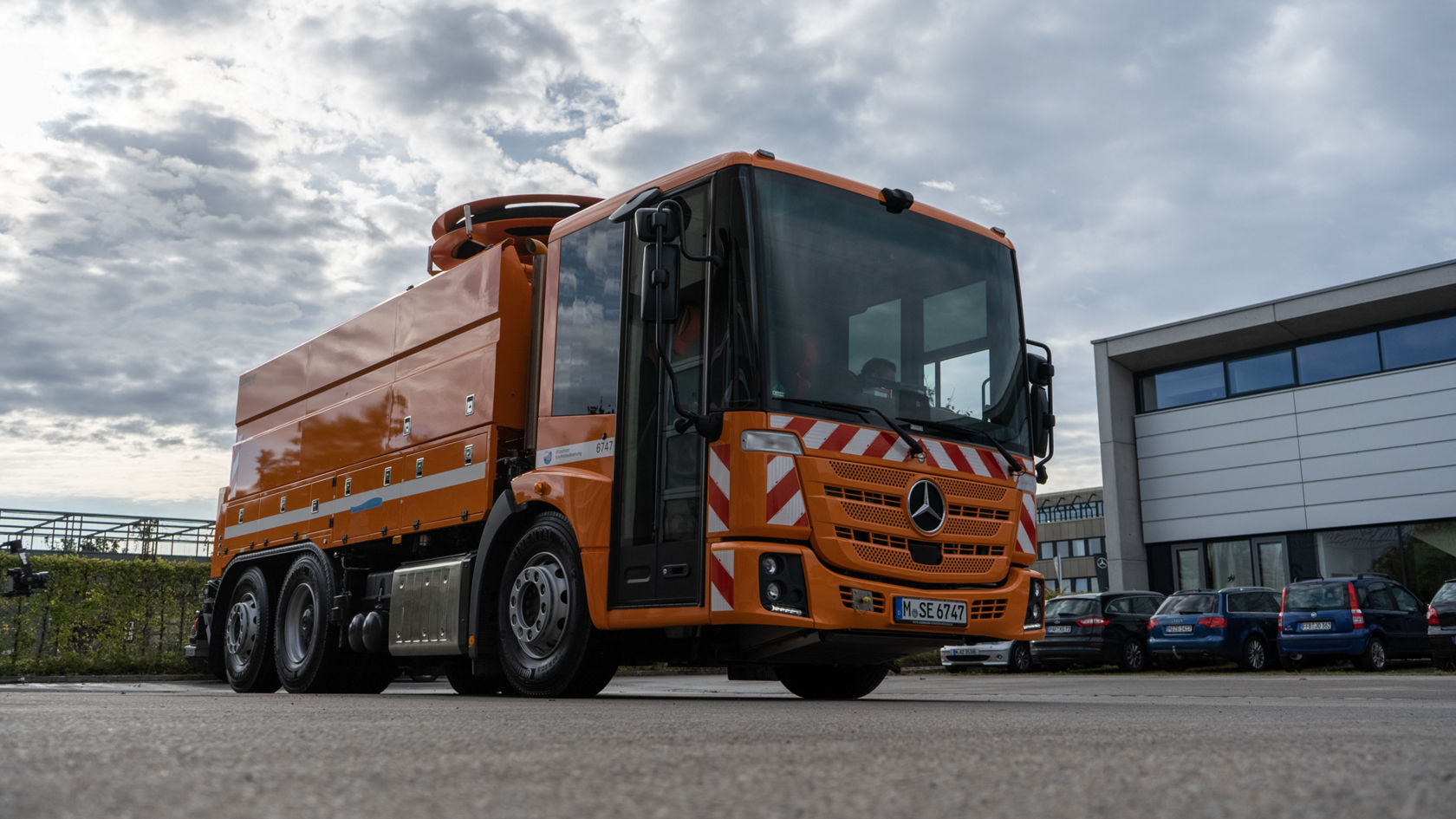 Mercedes-Benz Econic in leuchtendem Orange, mit einem großen Aufsatz auf dem Fahrzeugdach, steht auf einer Straße vor modernen Gebäuden.