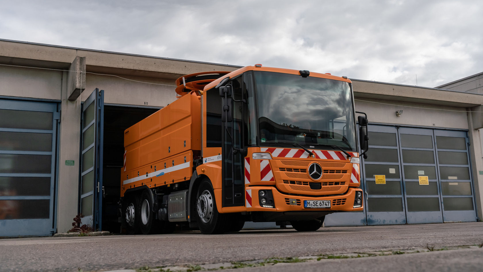 Ein orangefarbener Mercedes-Benz Econic-Fahrzeug verlässt eine Lagerhalle. Das Fahrzeug ist mit Warnstreifen ausgestattet und bereit für den Einsatz.