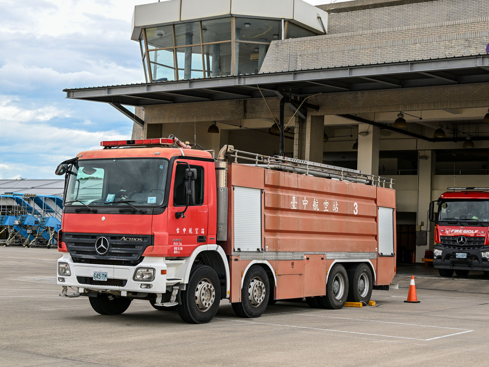 Ein rotes Feuerwehrfahrzeug der Marke Mercedes-Benz steht vor einem Feuerwehrgebäude. Im Hintergrund ist ein weiteres Feuerwehrfahrzeug sichtbar.