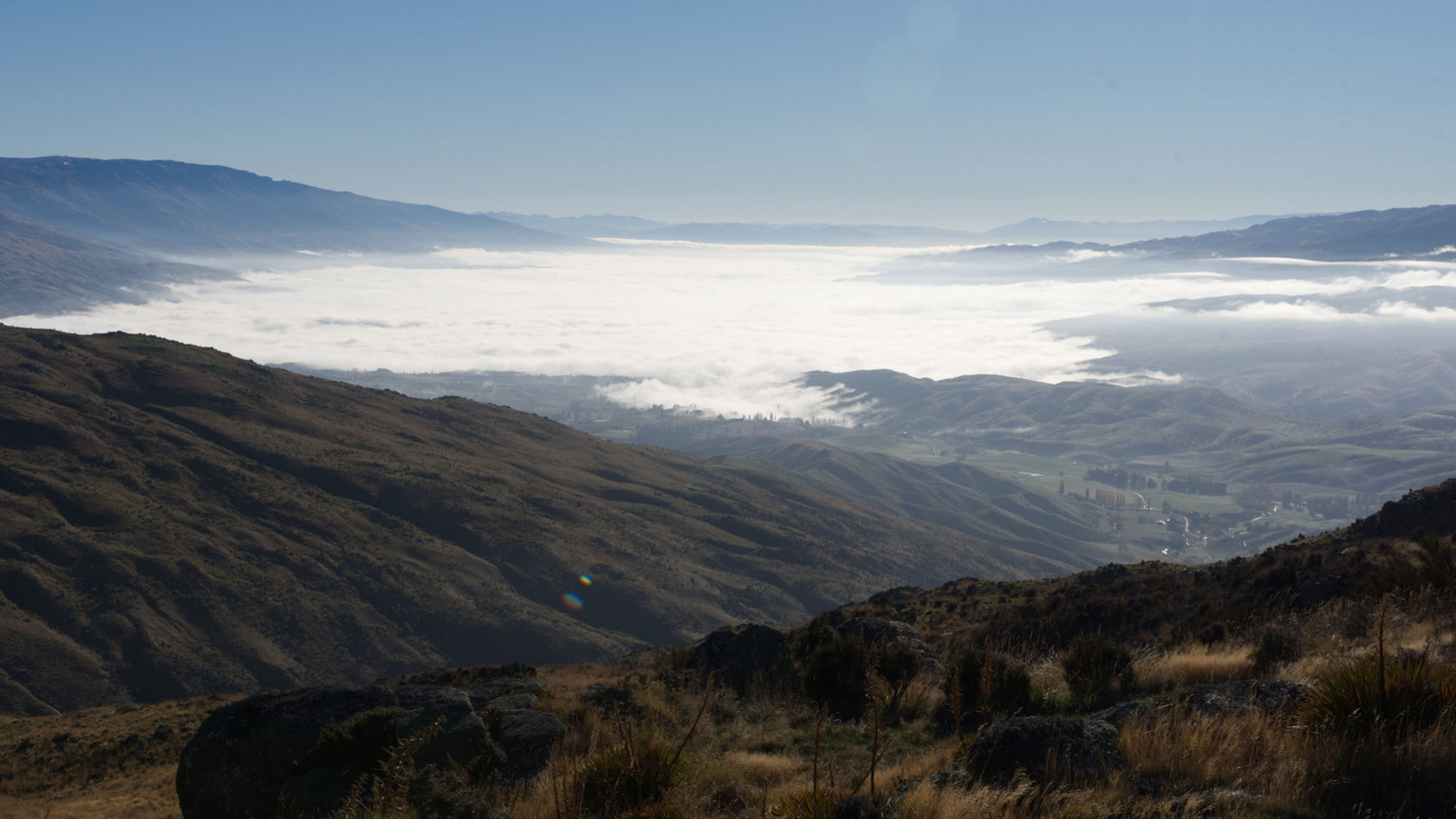 Blick auf einen nebligen Talboden, umgeben von Bergen, unter einem strahlend blauen Himmel mit der Sonne im Hintergrund. Ideal für Natur- und Landschaftsfotografie.