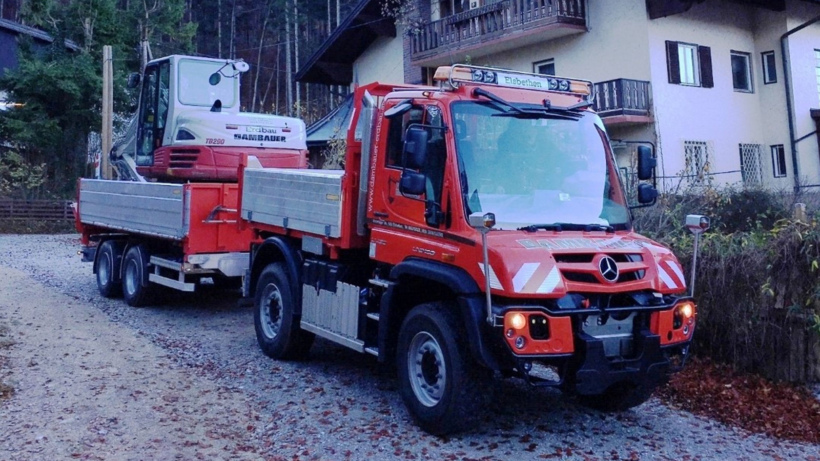 Ein rotes Mercedes-Benz Nutzfahrzeug zieht einen Anhänger mit einem kleinen Bagger auf einem Kiesweg, umgeben von Bäumen und einem Wohnhaus im Hintergrund.