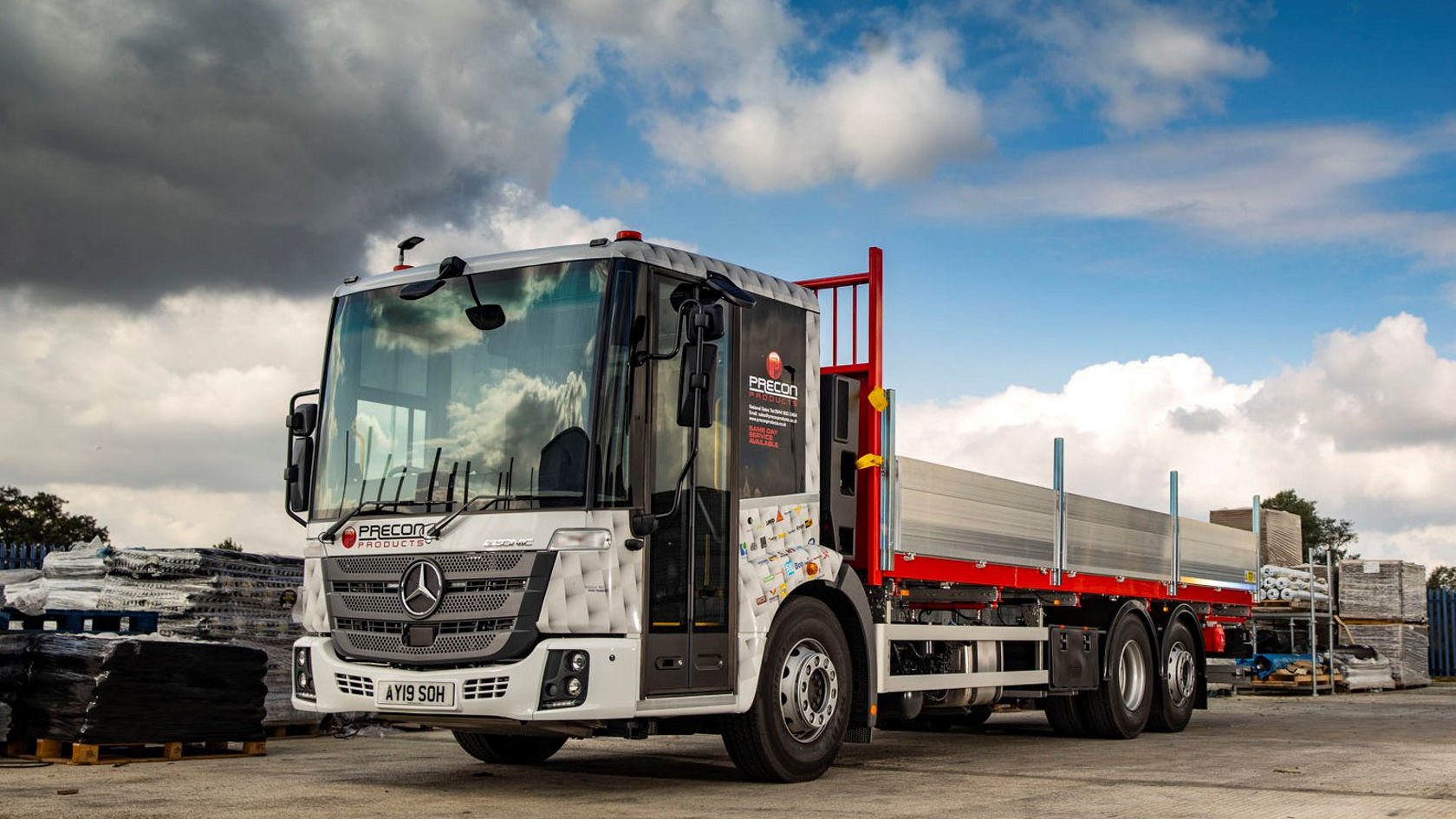 Mercedes-Benz Econic Lkw mit offene Ladefläche steht auf einem Baustellengelände, umgeben von Baustellenmaterialien, unter einem bewölkten Himmel.