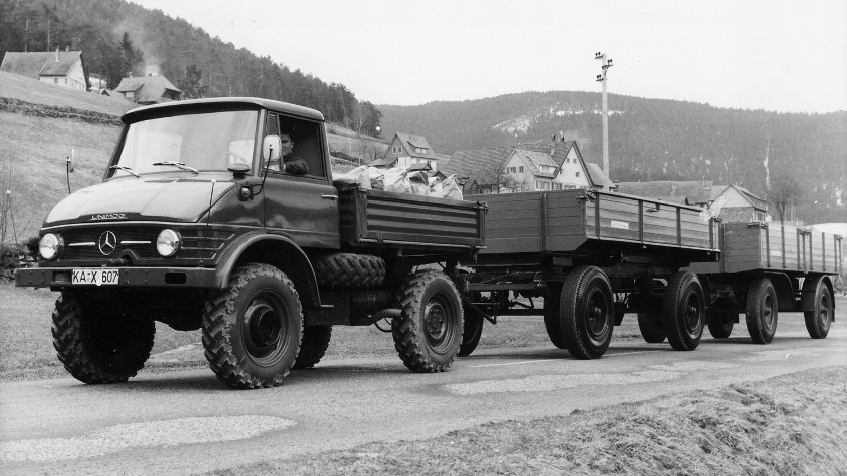 Een Mercedes-Benz Unimog met aanhangwagen staat op een weg, omgeven door het landelijke landschap, en dient als een veelzijdig bedrijfsvoertuig voor transporttaken.