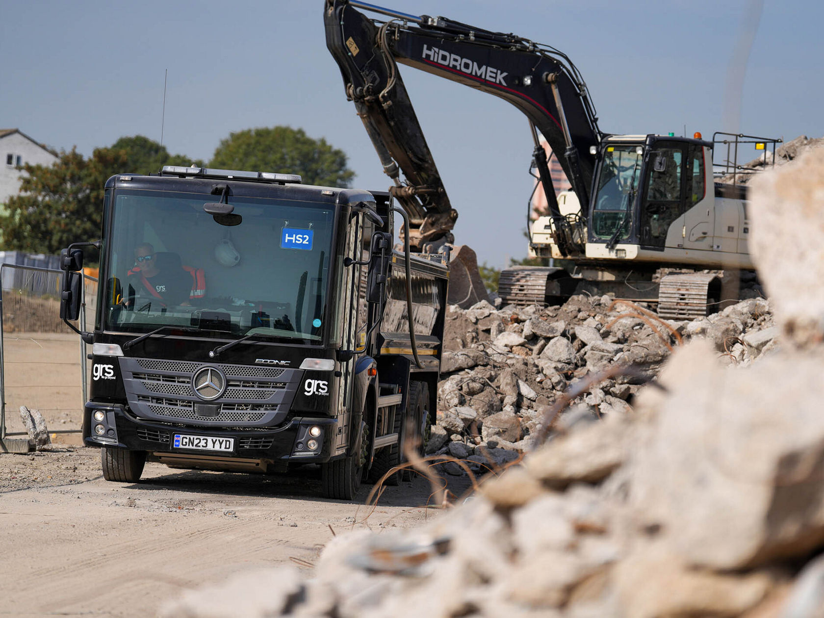 Ein schwarzer Mercedes-Benz Lkw steht auf einer Baustelle, während ein Bagger der Marke Hidromek hinter ihm mit Betonstücken arbeitet.