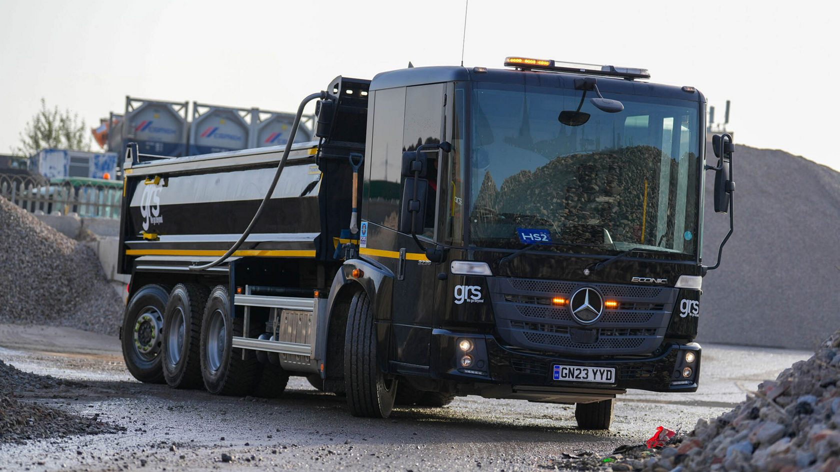 Ein schwarzer Mercedes-Benz Lkw steht auf einer Baustelle, umgeben von Kieshaufen. Der Lkw hat eine große Mulde und einen Anhänger für den Transport von Materialien.