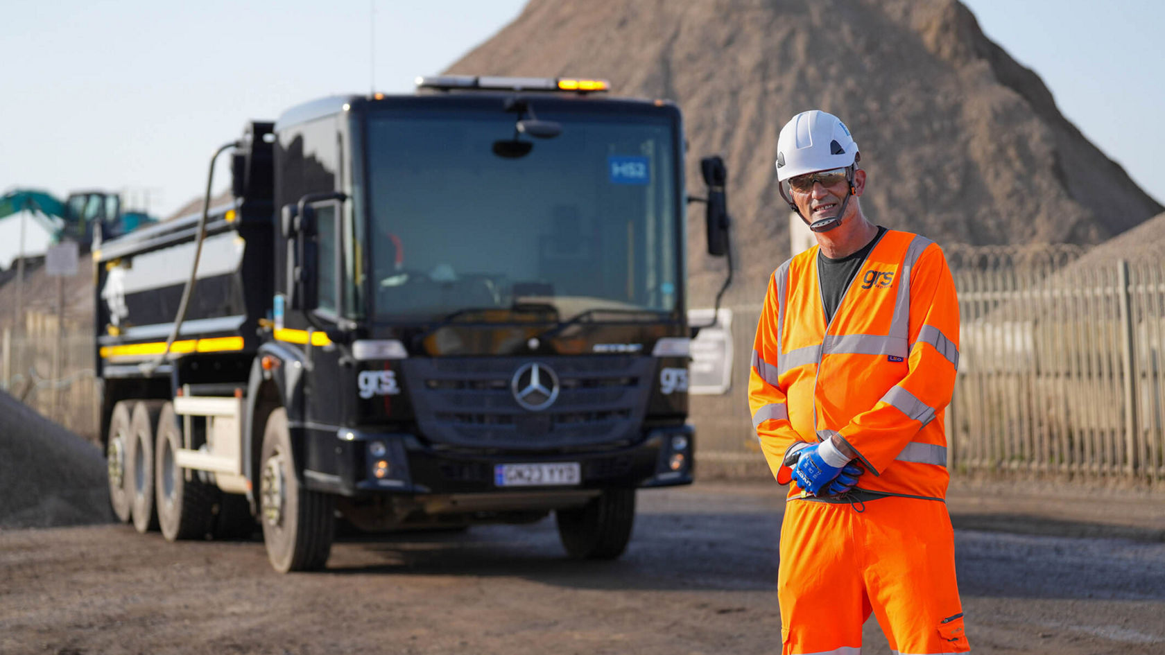 Ein Sicherheitsmitarbeiter in leuchtoranger Arbeitskleidung und Helm steht vor einem schwarzen Mercedes-Benz Lkw auf einem Baustellengelände mit Sandhaufen im Hintergrund.