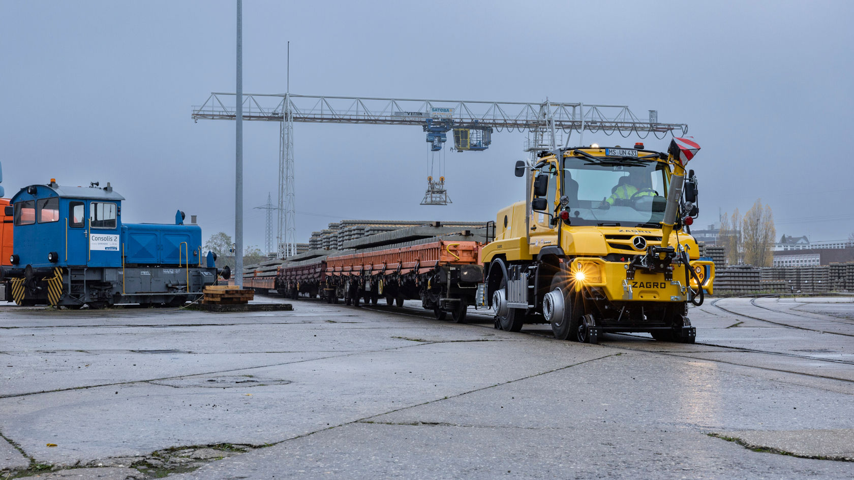 Ein Zweiwege Unimog fährt auf den Schienen eines Betriebsgeländes. Er zieht einen langen Anhänger mit Gleisschwellen.