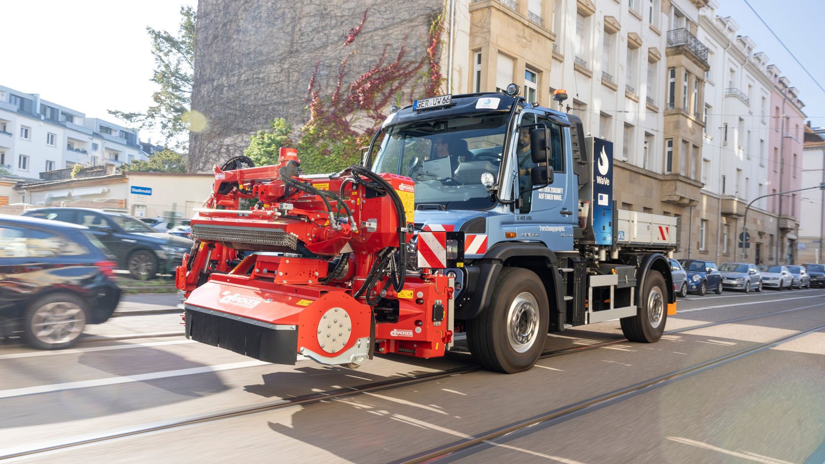 Ein Mercedes-Benz Unimog fährt auf einer Straße und ist mit einer Straßenreinigungsmaschine ausgestattet, die vorne montiert ist.