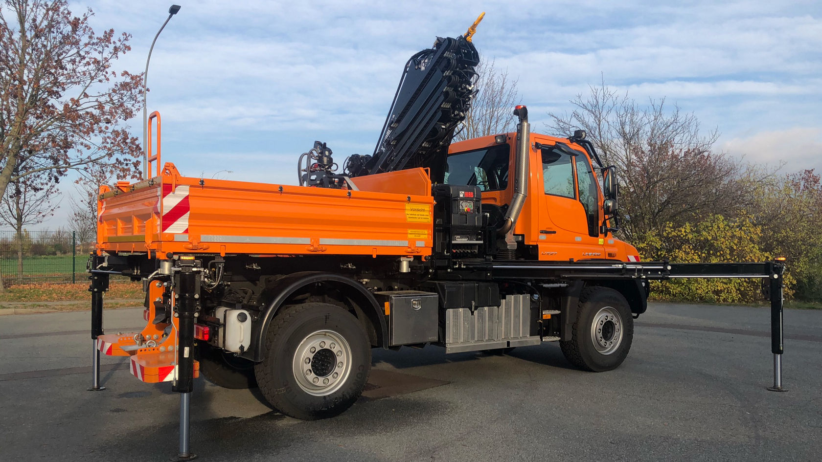 Een oranje Mercedes‑Benz Unimog met een vrachtwagenopbouw en een kraanarm op de laadvloer staat op een weg. De kraanarm is naar boven uitgeschoven.