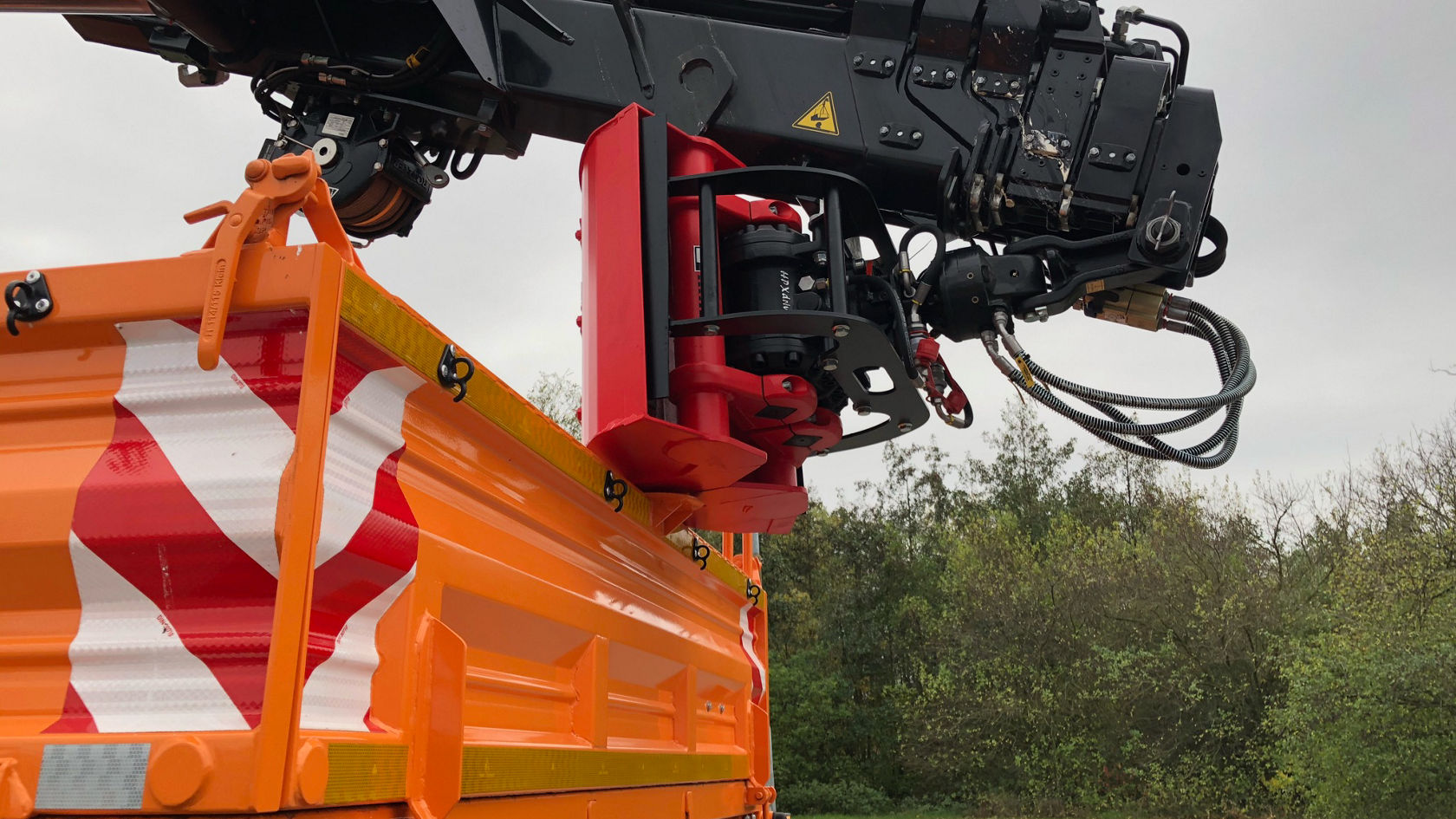 Een kraanarm met hydraulische slangen en besturingseenheden, gemonteerd op een oranje voertuigopbouw. De constructie is ontworpen voor laad- en hijswerkzaamheden.
