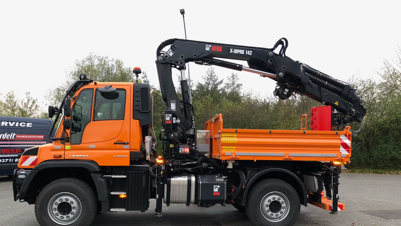 Een Mercedes-Benz Unimog met een kraanopbouw, een oranje opbouw en een container op de achtergrond, geparkeerd op een verharde plaats.