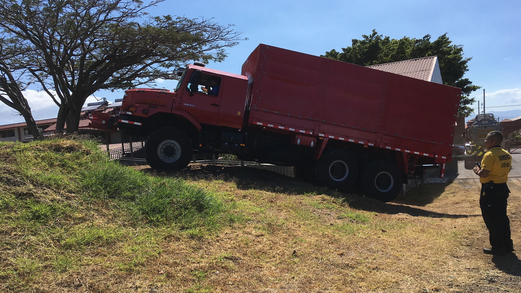Un camion Mercedes-Benz rouge passe en biais devant une colline, avec un arbre en arrière-plan. Le plateau de chargement du camion est positionné à une hauteur inhabituellement élevée.