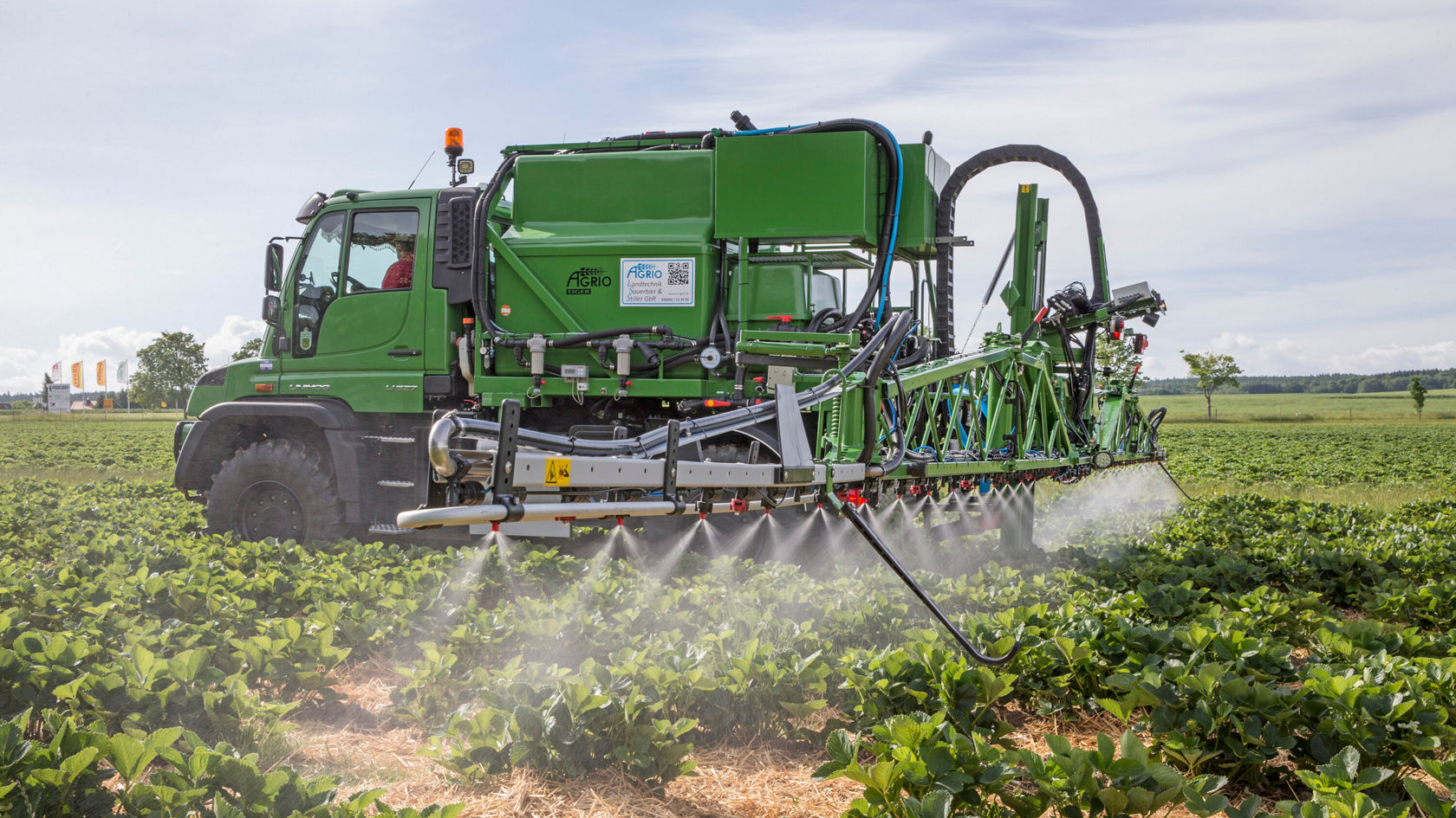A Unimog drives across a field with plants. The vehicle sprays a fine mist onto the plants.