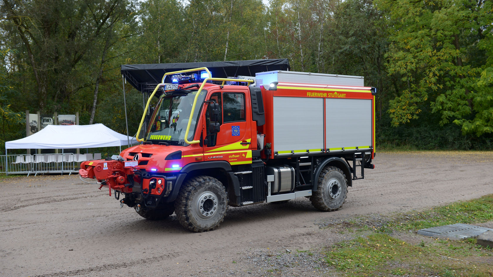 Ein rotes Einsatzfahrzeug der Feuerwehr Stuttgart von Mercedes-Benz, ausgestattet mit Blaulicht, steht auf einem unbefestigten Gelände.