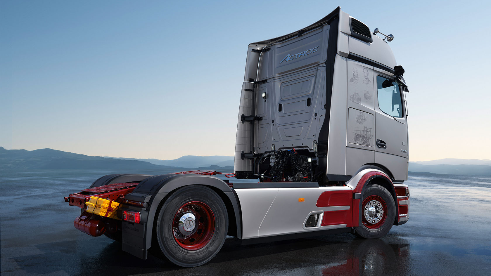 從後方可見一輛 Mercedes-Benz Actros 半掛牽引車，採用 Legacy Silver Metallic 車色，輪拱與輪圈飾以樞機紅色點綴。駕駛室後側醒目地標有 'ACTROS' 字樣，車側則繪有歷史卡車素描。車輛停放在反光的瀝青路面上，背景為群山景致。