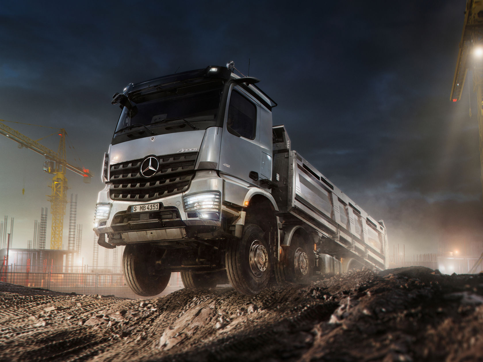 Ein silberner Mercedes-Benz Baustellen-Lkw fährt bei Nacht über unebenes Gelände auf einer Baustelle. Im Hintergrund sind Baukräne zu sehen.