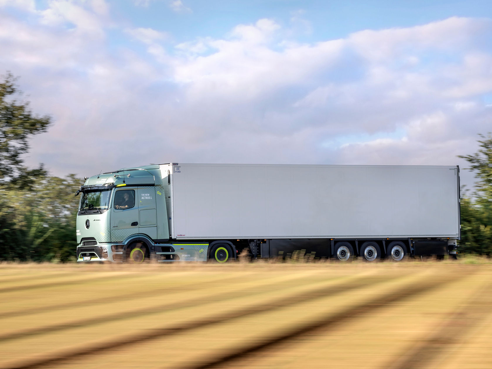 Ein hellgrüner Mercedes-Benz Lkw mit einem weißen Auflieger fährt schnell auf einer Landstraße. Der Hintergrund ist durch die Bewegung verschwommen.