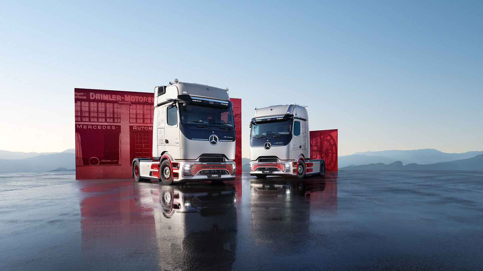 兩輛 Mercedes-Benz 卡車（Actros L 與 eActros）採用 Legacy Silver Metallic 車色，並以鮮明的紅色點綴，並排停放在潮濕、鏡面反射的地面上，背景為山景及展示戴姆勒歷史題材的紅色展示板。