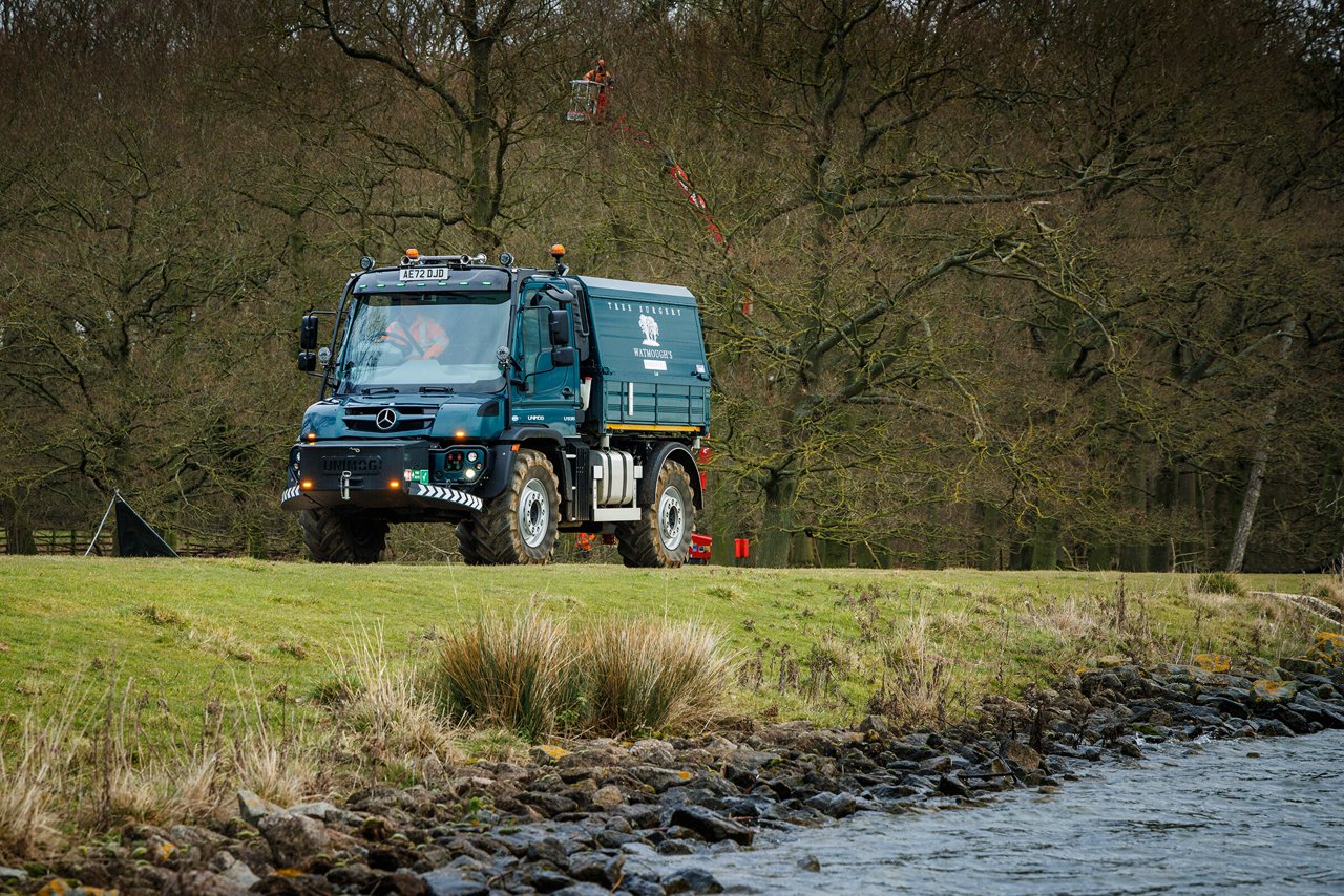 Ein geländetauglicher Mercedes-Benz Unimog U 535 steht auf einer Wiese am Ufer eines Gewässers, umgeben von Bäumen. 