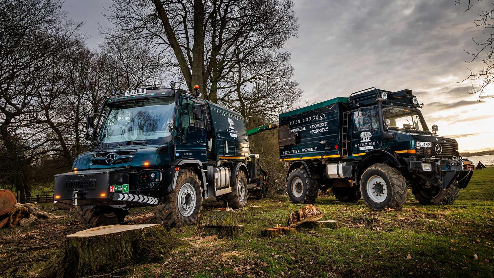 Zwei Mercedes-Benz Unimog stehen in einem Waldgebiet, umgeben von gefällten Baumstümpfen. Die Fahrzeuge sind in dunklen Farben lackiert und mit Logos versehen.