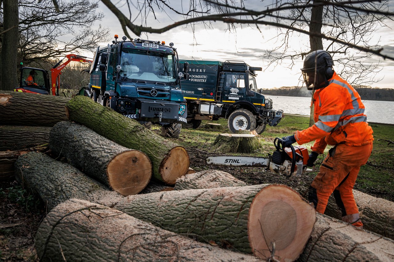 Ein Arbeiter in Sicherheitskleidung benutzt eine Motorsäge, um Baumstämme zu bearbeiten. Im Hintergrund sind zwei Mercedes-Benz Unimog zu sehen.