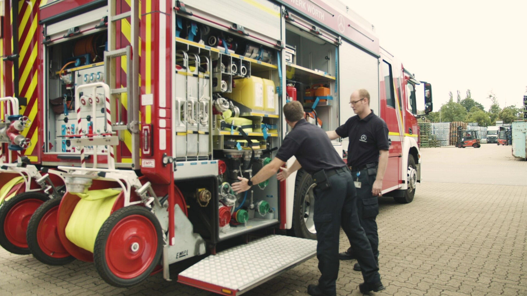 Un véhicule de pompiers Mercedes‑Benz rouge, avec l'arrière ouvert et équipé de tuyaux et de matériel d'extinction, est stationné tandis qu'un pompier se tient à côté.
