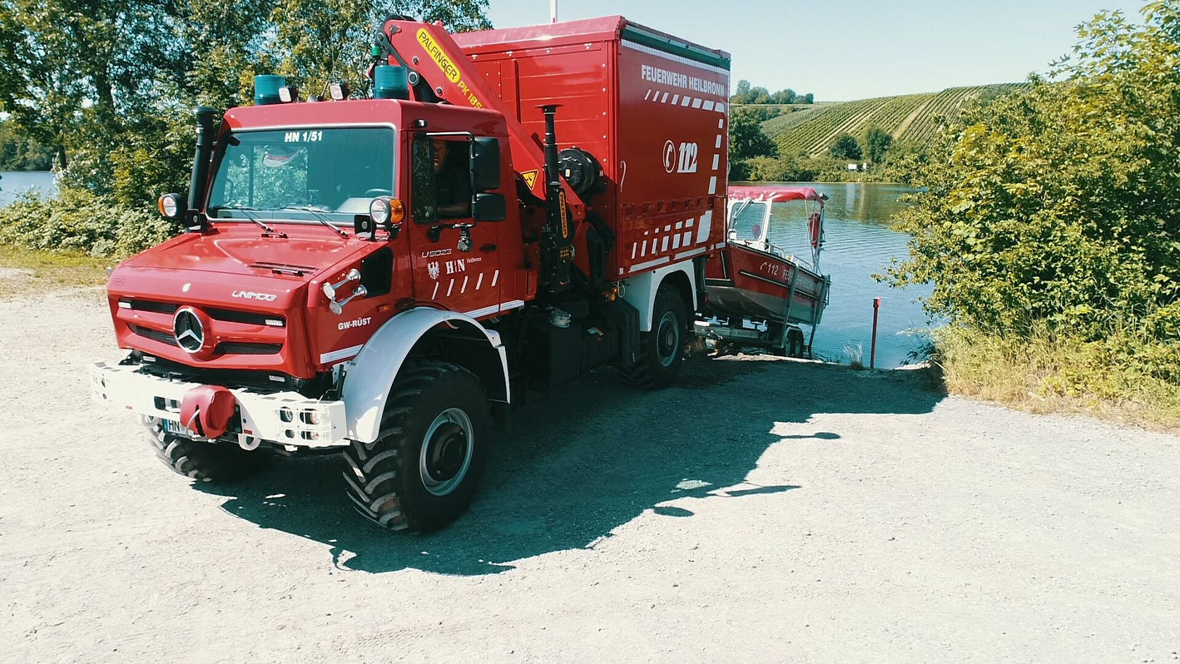 Un camion de pompiers Mercedes‑Benz rouge est stationné au bord d'un plan d'eau, tandis qu'un pompier en tenue d'intervention descend du véhicule. En arrière‑plan, un bateau est visible sur la rive.