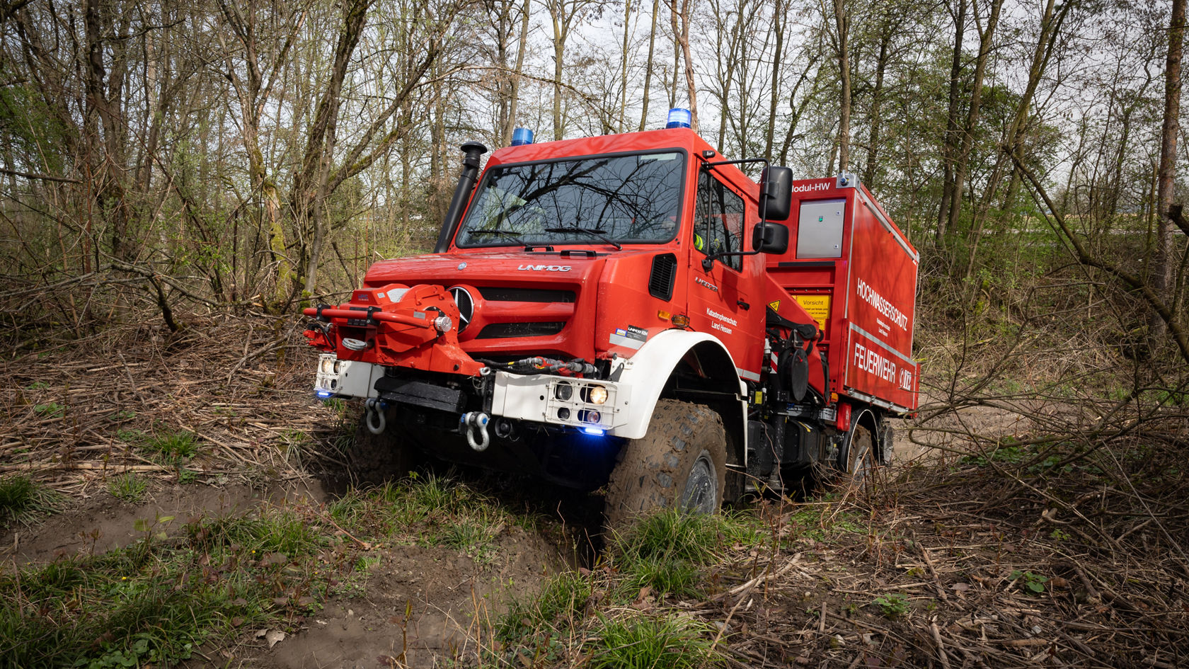 Un véhicule de lutte contre les incendies Mercedes‑Benz rouge se trouve sur un terrain accidenté en forêt et est équipé d'un gyrophare et d'un treuil, prêt à intervenir.
