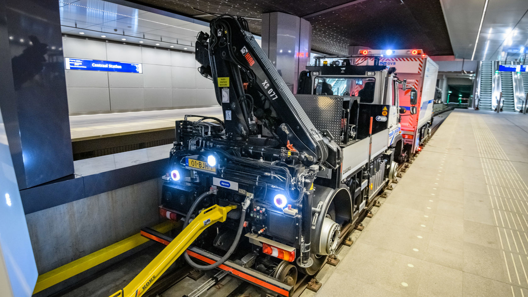 Ein Mercedes-Benz Unimog steht auf Gleisen in einer U-Bahn-Station, ausgestattet mit einem Kranarm und Fahrzeuganhänger, bereit für Wartungsarbeiten.