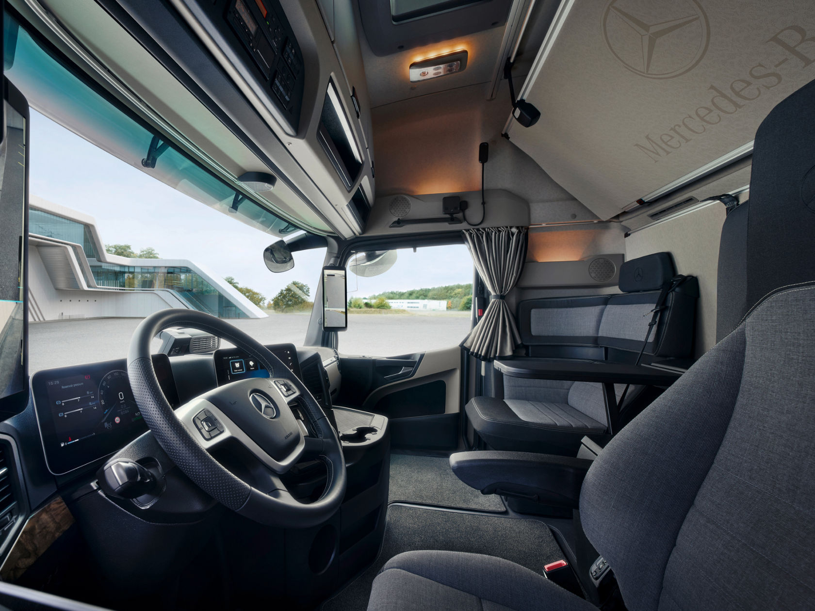 L'image montre l'intérieur spacieux et confortable d'un camion Mercedes-Benz moderne. On voit le volant, les écrans numériques et l'espace de vie avec une table et des sièges, ce qui souligne la fonctionnalité et le confort pour les longs trajets.