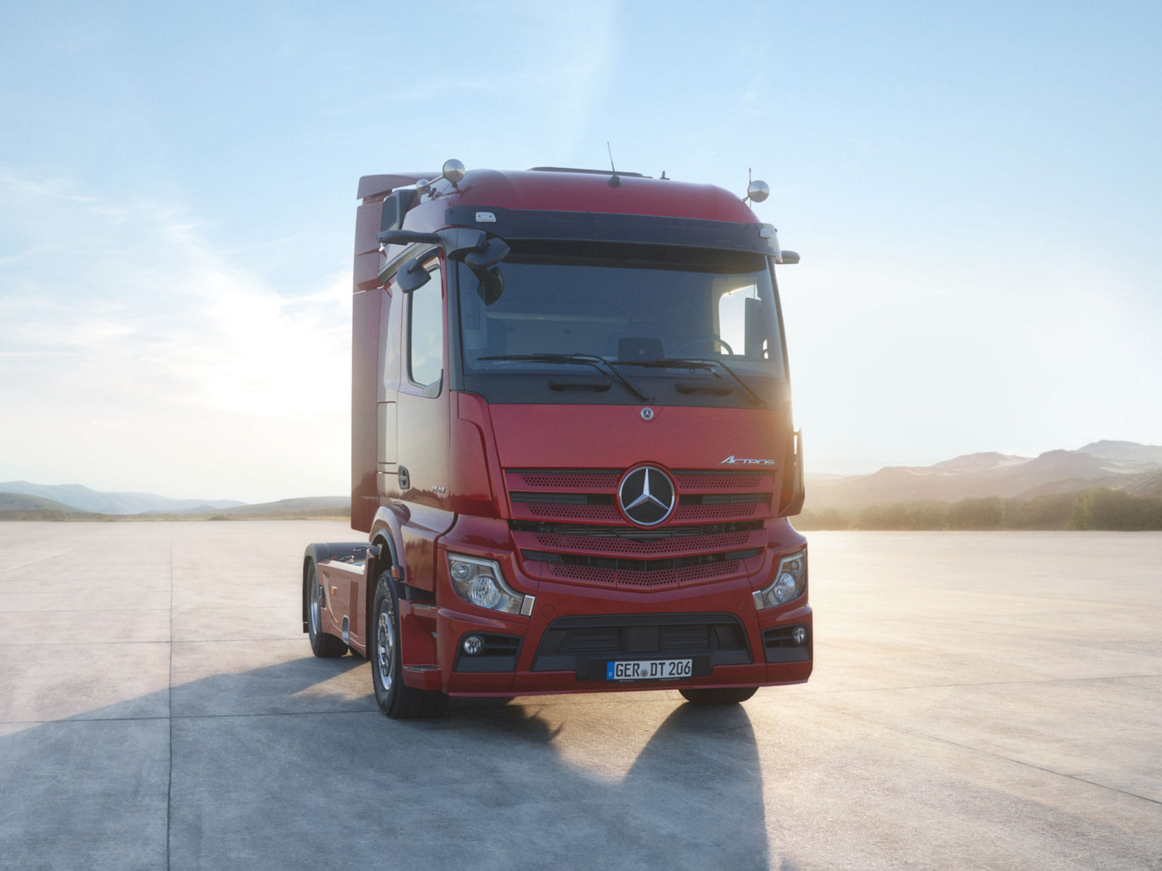 Na obrázku je červený tahač Actros, který stojí na betonové ploše. V pozadí jsou vidět hory a modrá obloha. Slunce svítí a odráží stín nákladního vozidla na zem. 
