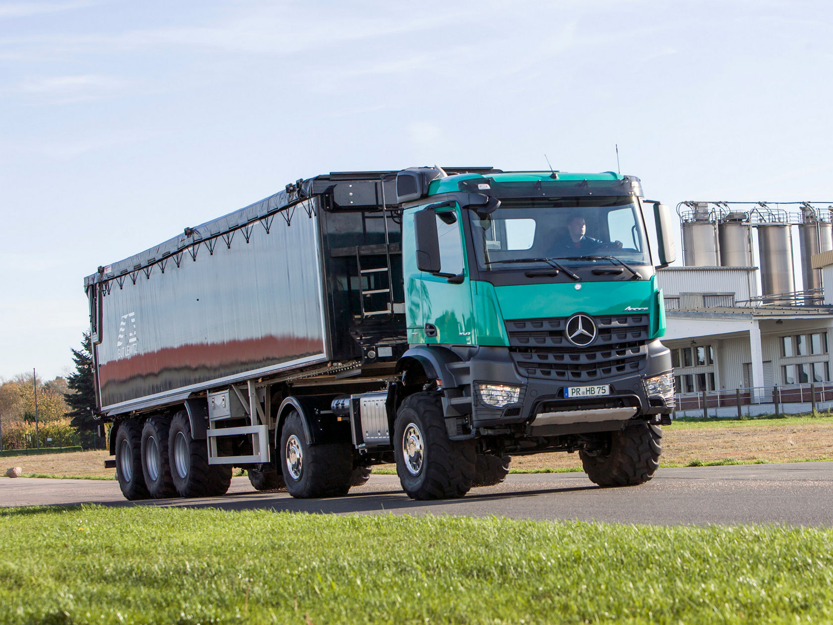 Картината показва зелен Arocs с черно ремарке, който се движи по път. На заден фон се виждат сграда и няколко силоза. Небето е синьо и има няколко облака.