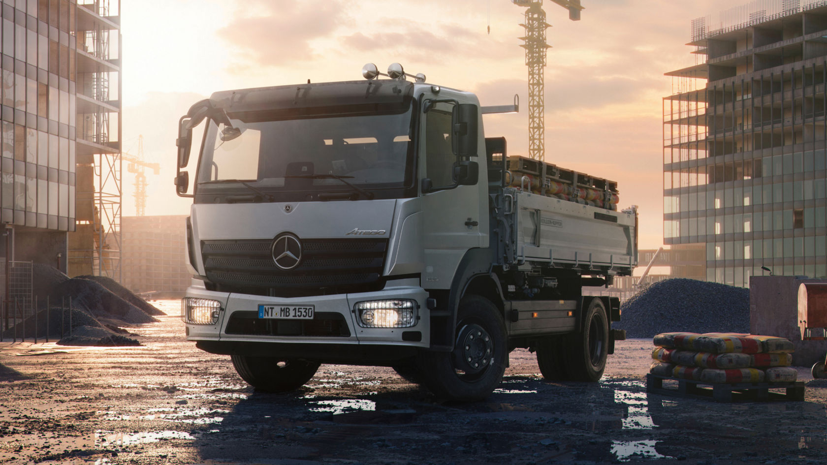 Картината показва бял Atego на строителна площадка. На заден фон се виждат недовършени сгради и строителен кран. Почвата е кална и има локви. На товарната повърхност на камиона има палети със строителни материали. Небето е облачно и слънцето свети през облаците.