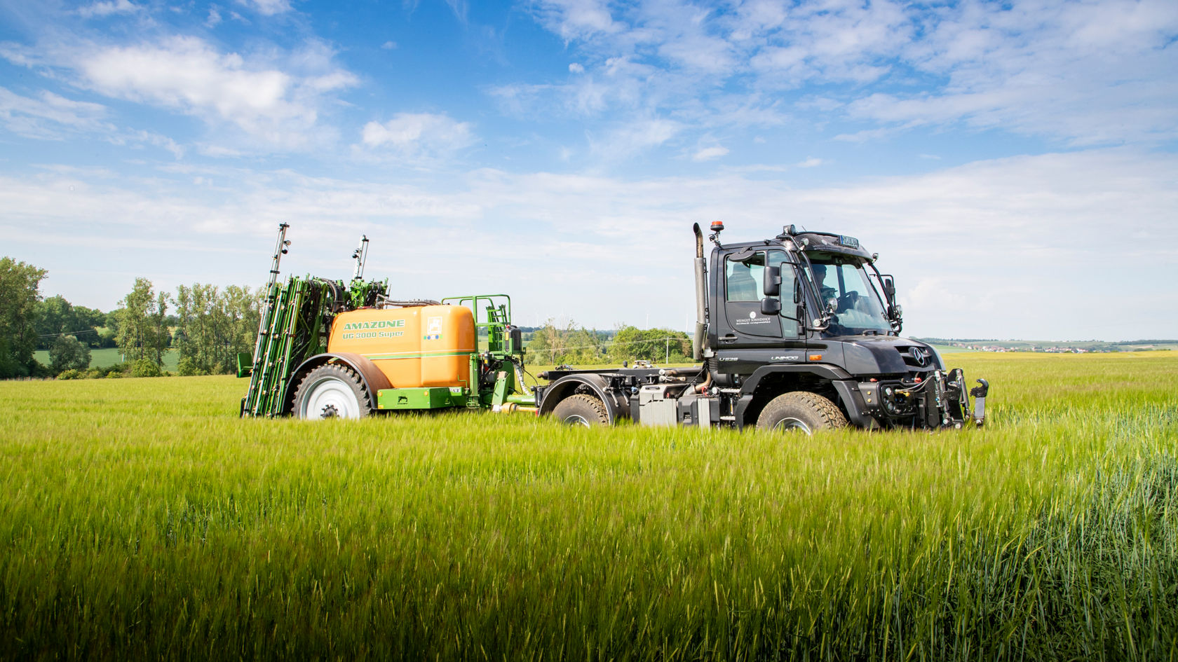 A black Unimog driving across a green grain field. It is pulling an orange Amazone field sprayer. In the background is a blue sky with white clouds with more fields and trees on the horizon.