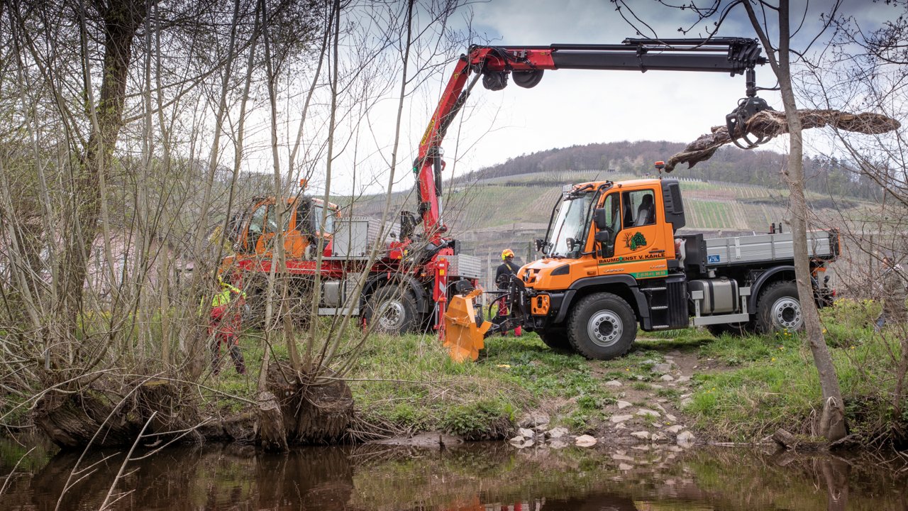 An orange Unimog with a crane body standing on the banks of a river. The crane is lifting a tree trunk. In the background are fields, two other workers and another similarly equipped truck.