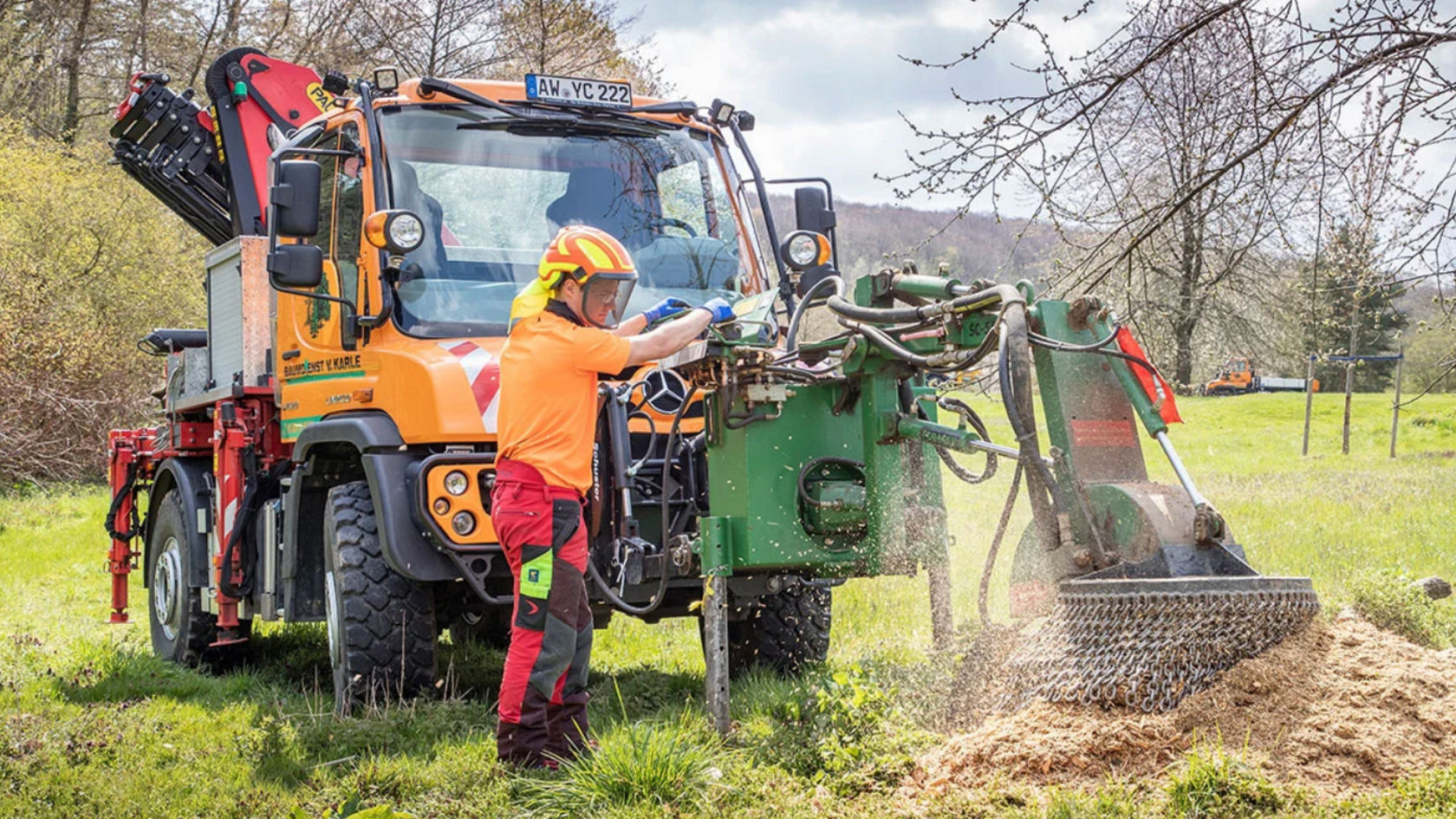 An orange Unimog standing in a meadow. A person in work clothes is operating a wood chipper at the front of the truck, from which wood chips emerge. Another truck can be seen in the background.