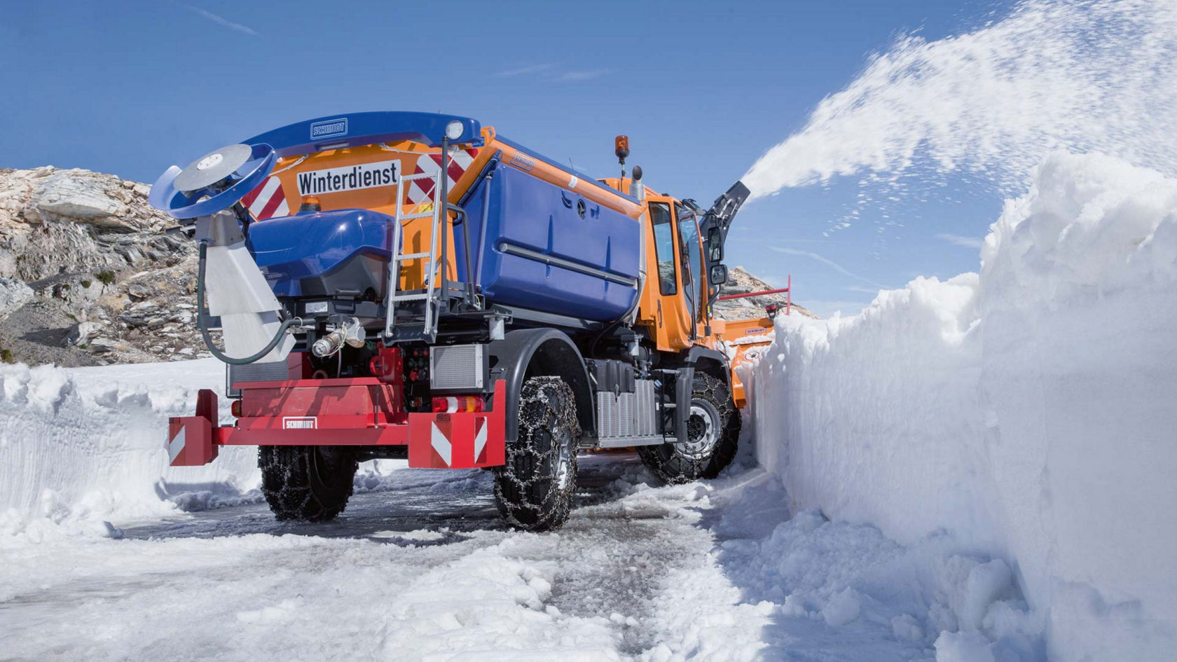 The image shows an orange-and-blue Unimog with a snow plough and snow blower to the rear. The truck has snow chains on the wheels and is clearing a snowy road. The snow blower is throwing the snow to the side. There is lots of snow and a rock in the background.