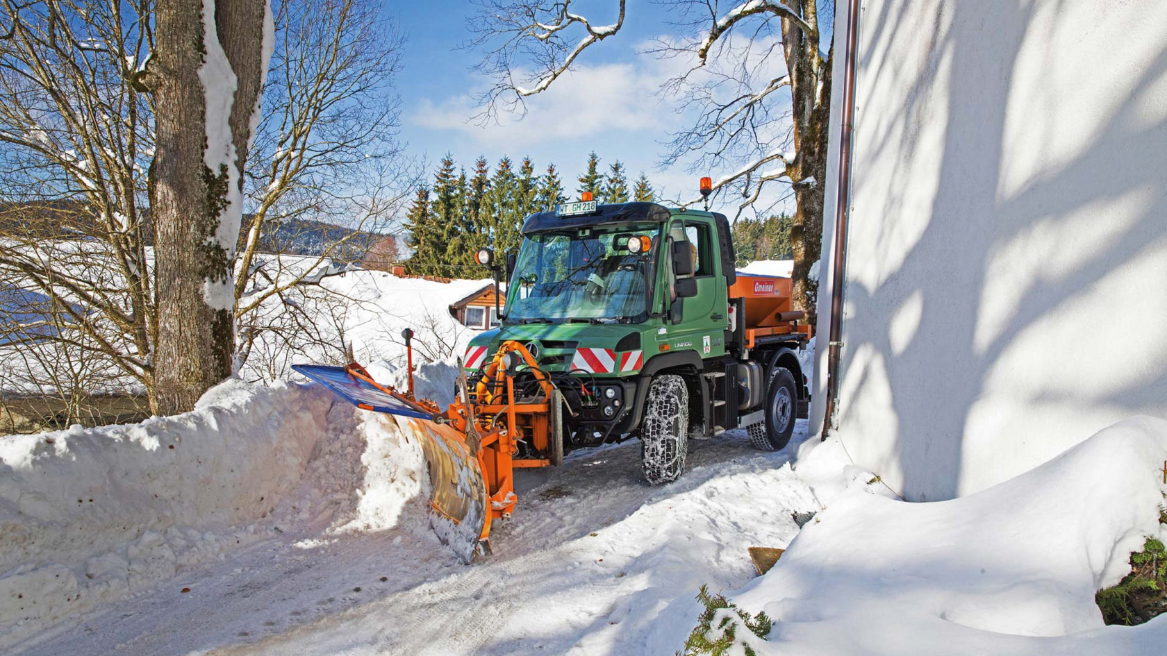 A green Unimog with an orange snow plough clearing snow from a road in a snowy village. Snow chains are fitted to the tyres. It is driving past a snow-covered wooden hut and trees. Lots of snow can be seen in the background and the sky is blue. 