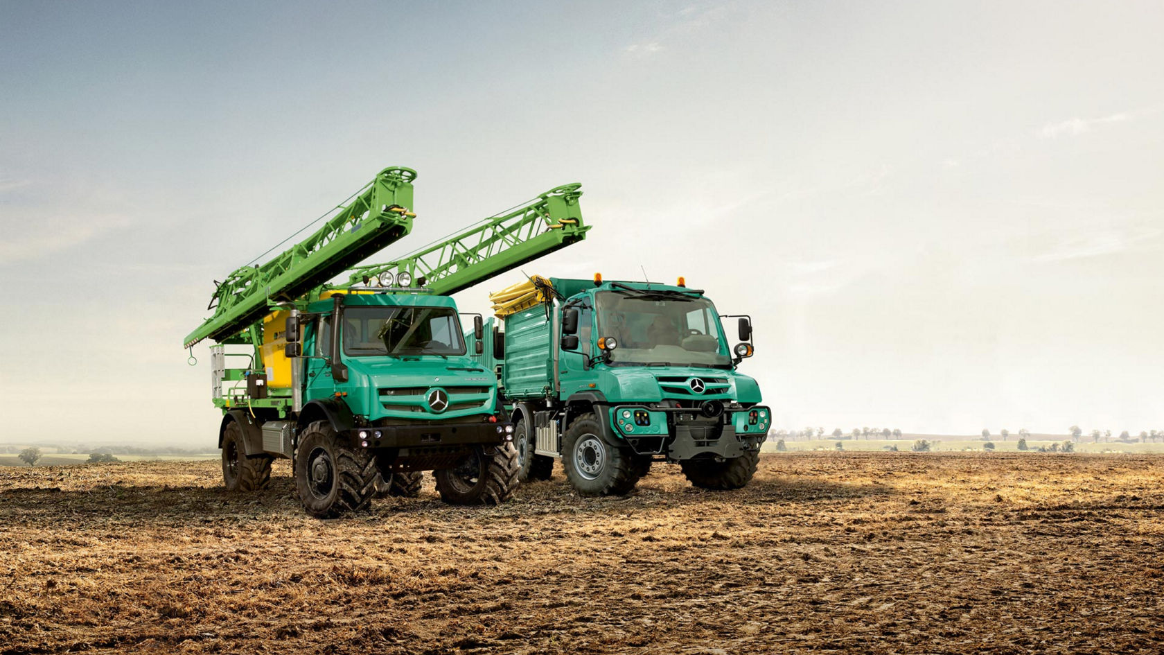 Two turquoise trucks (Unimog and Zetros) standing in a field. The truck on the left has extended a large, light green spraying rod. The truck on the right has a green tip box. A landscape with trees can be seen in the background.