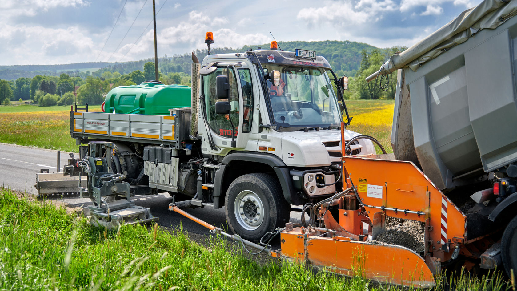 A white Unimog spreading grit on a roadside next to a meadow with yellow flowers. An orange grit spreader and a green water tank are mounted on the truck. A second truck with a tipper vehicle is standing next to it, delivering the grit.