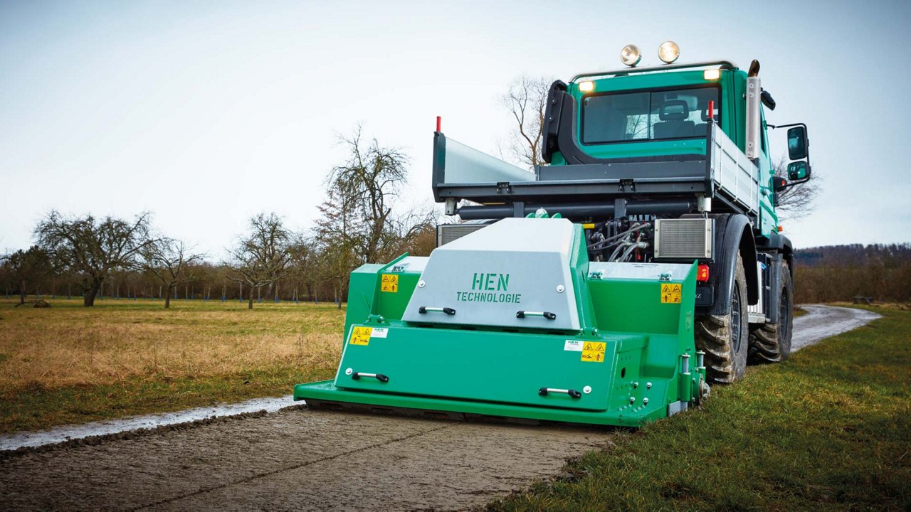 A green Unimog with a tipper vehicle pulling a green ground miller behind it. It is working on an unpaved path surrounded by meadows and trees. The miller says “HIEN Technology”.