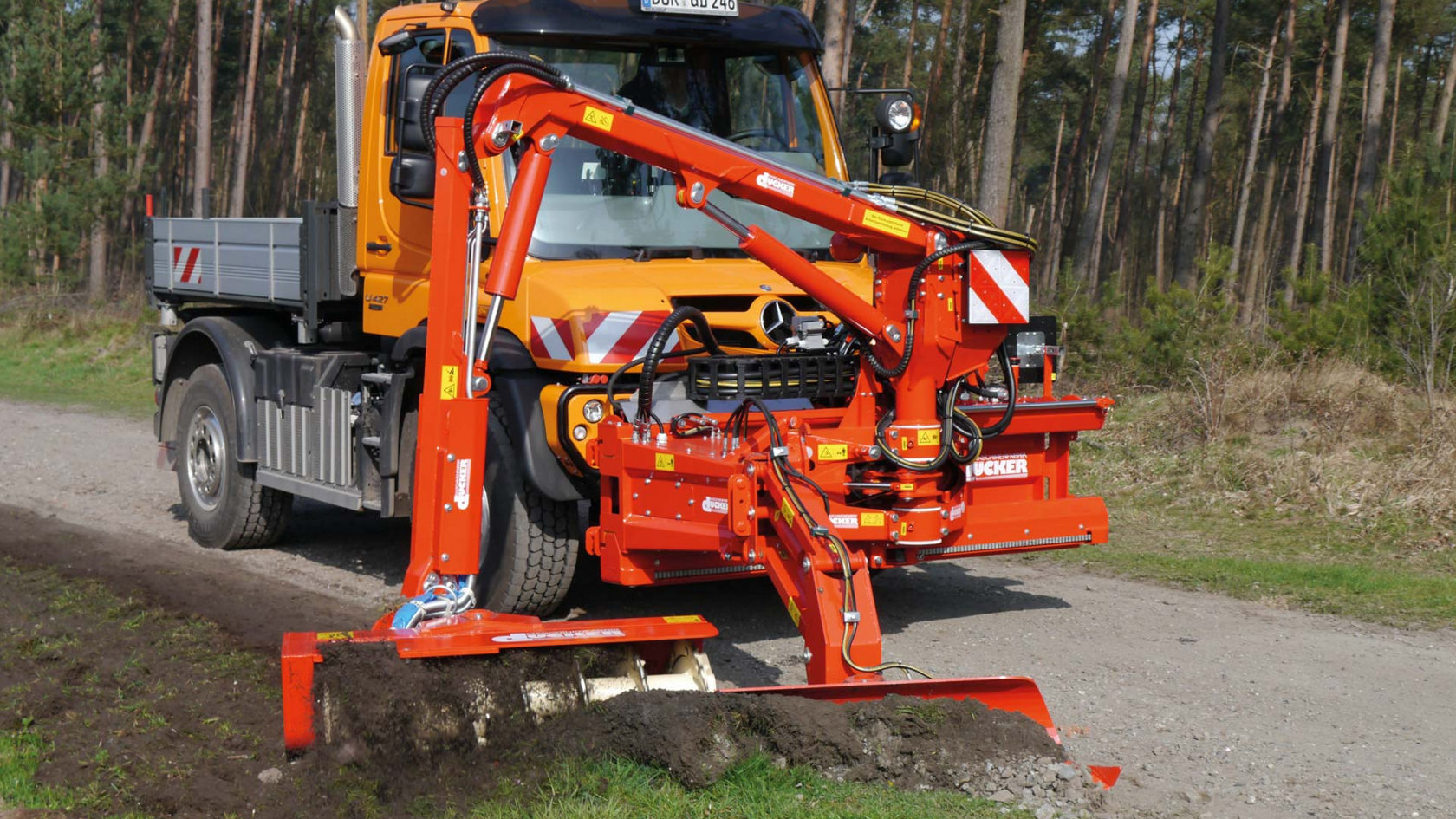 An orange Unimog with a loading area and crane standing on a forest path. An orange mower is attached to the crane, which is working on the roadside ditch.