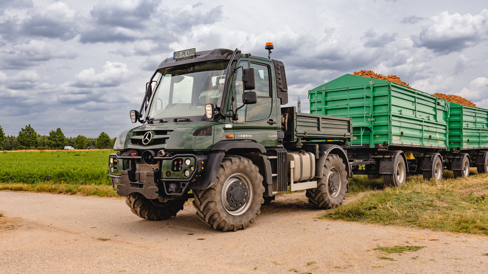 Grüner Unimog, der zwei grüner Anhänger auf einem Feld zieht.