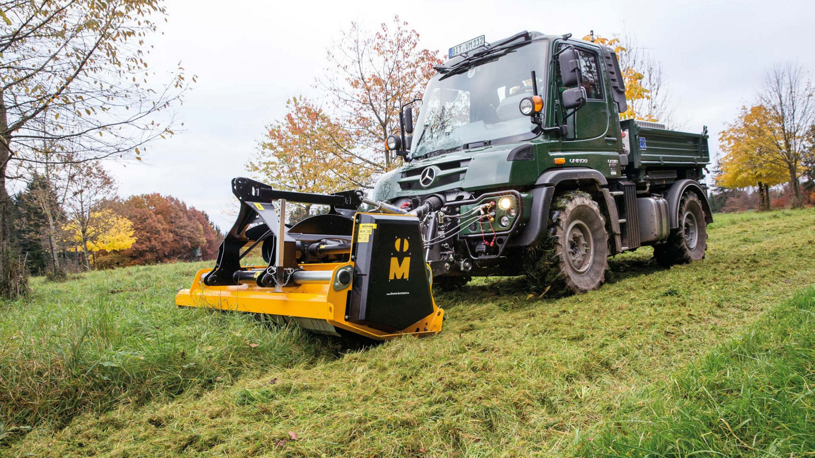 A dark green Unimog with a loading area mowing a meadow with a yellow mulcher. The truck is driving on the grass and the mower is mounted on the front of the truck. Trees are visible in the background.